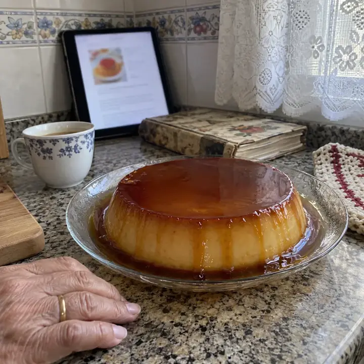 Flan de huevo recién desmoldado con caramelo en un plato de cristal, con una tablet apoyada en los azulejos